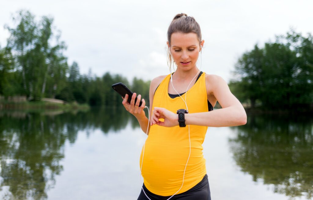 Pregnant woman using fitness app on smart watch and smartphone during exercises outdoor